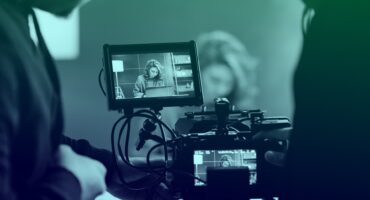 Photo of a video camera filming a scene in the background. We are looking into the camera as if we are operating it, and the background scene is slightly out of focus.