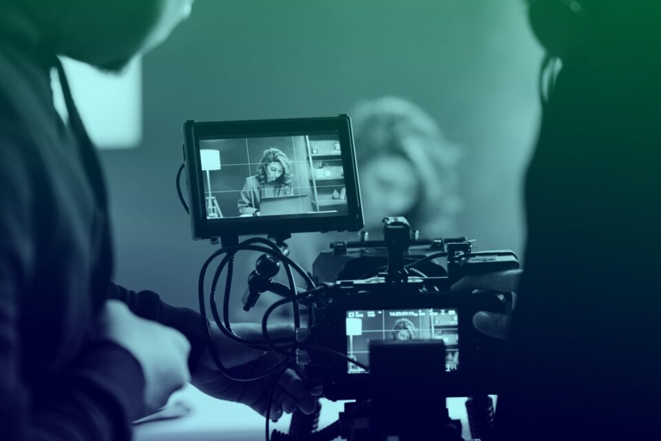 Photo of a video camera filming a scene in the background. We are looking into the camera as if we are operating it, and the background scene is slightly out of focus.